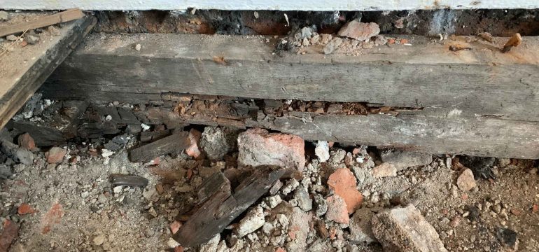 Exposed section of a damaged wooden beam with visible rot, surrounded by broken bricks, rubble, and debris near a white wall—an example of rotting wood in house structures.