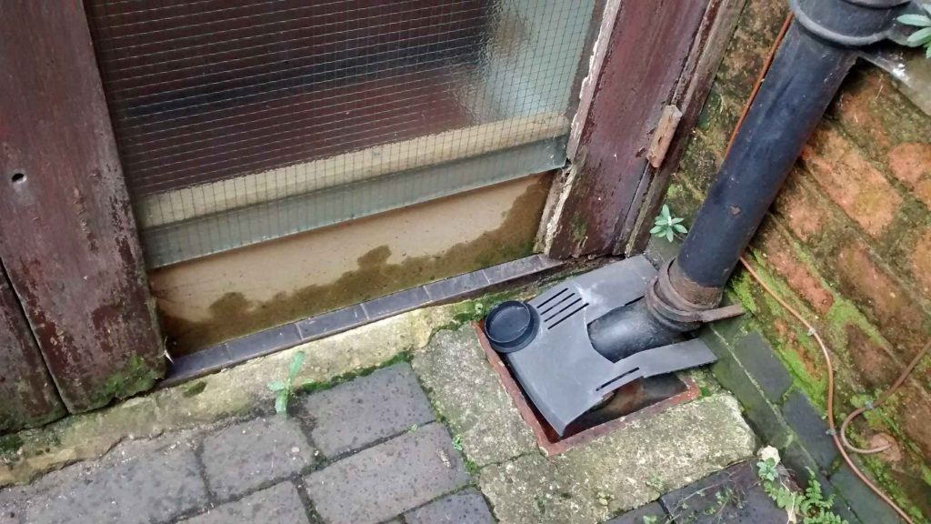 A black drainpipe connects to a drain in the ground near an old wooden door with a glass panel.