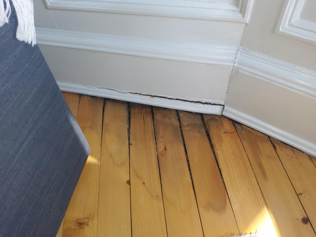 A close-up of a corner in a room with wooden floorboards and white baseboards, showing a large gap where the floor meets the wall. Part of a dark-coloured sofa is visible on the left.
